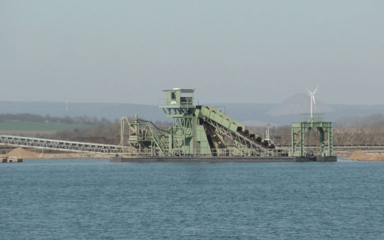 Eine schwimmende Förder- und Gewinnungsanlage liegt auf dem Wasser am Standort Ditfurt. Grüne Stahlkonstruktionen, Förderbänder, Plattformen und die umliegende Uferlandschaft prägen die industrielle Szene.