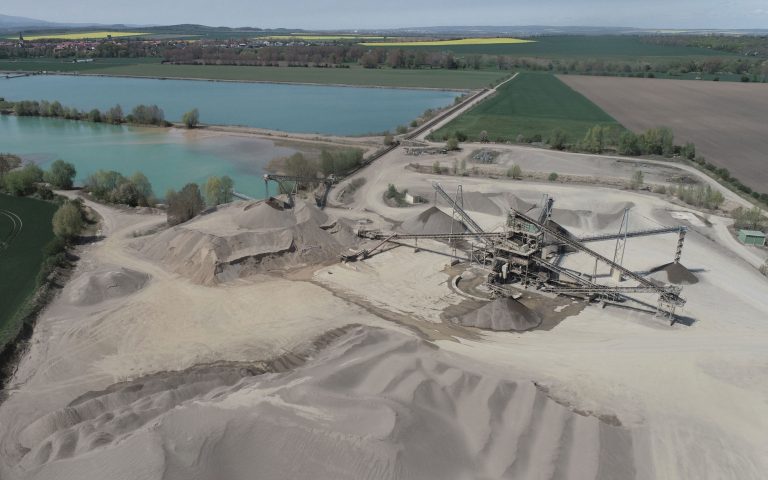 Ein großes Kiesabbaugebiet mit Sand- und Kieshalden, Fördertechnik und breiten Fahrwegen liegt direkt an türkisblauen Seen. Im Hintergrund sind Felder, Bäume und eine Ortschaft zu sehen.