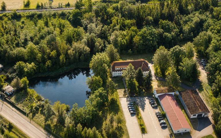Eine Luftaufnahme zeigt das Verwaltungsgebäude mit Nebengebäuden, Parkflächen und einem angrenzenden Teich, eingebettet in eine grüne, baumbestandene Umgebung.