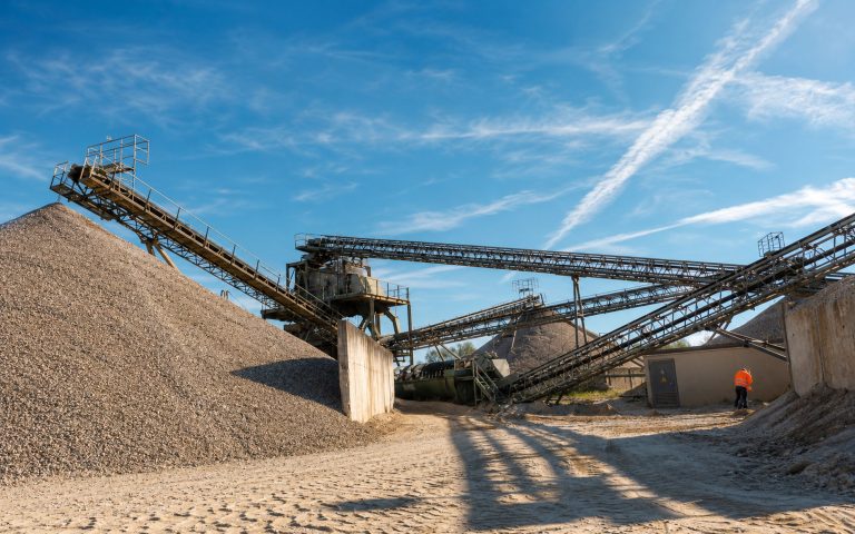 Mehrere Förderbänder überspannen ein Kieswerk mit großen Materialhalden und sandigem Fahrweg. Ein Mitarbeiter in Warnkleidung steht am Rand der Anlage unter klarem Himmel.