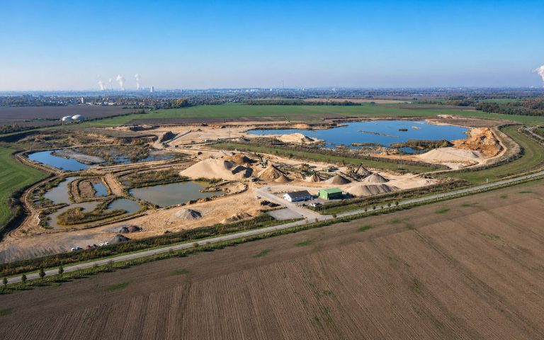 Eine weite Luftaufnahme zeigt ein großes Kies- und Sandabbaugebiet mit Wasserflächen, Materialhalden, Fördertechnik, Betriebsgebäuden und umliegenden Feldern.