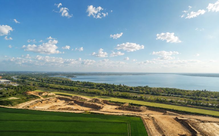 Eine Luftaufnahme zeigt ein weitläufiges Kiesabbaugebiet mit Sandflächen, Fahrwegen und Abbaukanten. Im Hintergrund liegt ein großer See mit grünen Uferbereichen und weiter Landschaft.