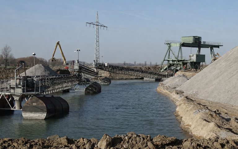 Eine industrielle Förder- und Gewinnungsanlage liegt an einem Wasserkanal. Förderbänder, Plattformen, Kieshalden und Maschinen prägen die Arbeitsfläche am Ufer.