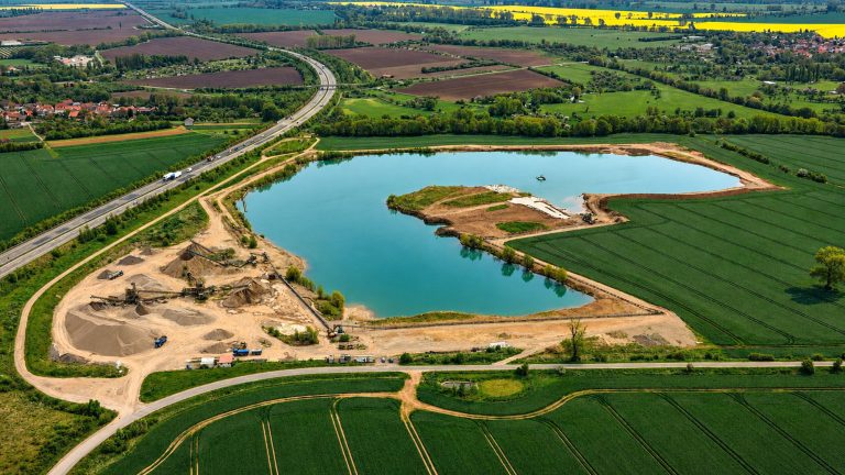 Das Bild zeigt einen gelben Radlader in einer offenen Kiesgrube bei der Materialaufnahme an einer steilen Abbauwand. Sichtbar sind die freigelegten Erd- und Kiesschichten, die große Schaufel des Fahrzeugs und die weitläufige Arbeitsfläche des Abbaugebiets. Die Szene vermittelt den laufenden Rohstoffabbau in einer trockenen, offenen Grubenlandschaft.