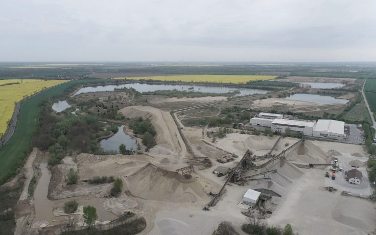 Eine Luftaufnahme zeigt ein weitläufiges Kieswerk mit Materialhalden, Förderanlagen, Betriebsflächen und umliegenden Seen, Feldern und Windkraftanlagen.