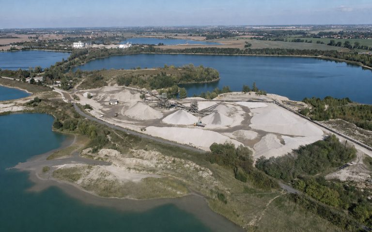 Eine Luftaufnahme zeigt ein Kieswerk mit hellen Materialhalden, Förderanlagen und Fahrwegen, eingebettet zwischen mehreren Seen, Uferbereichen und grüner Landschaft.