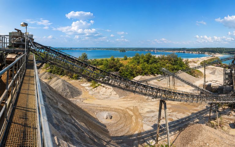 Ein weites Panorama zeigt ein Kieswerk mit langen Förderbändern, Materialhalden und Aufbereitungsanlagen. Im Hintergrund liegen ein See, Waldflächen und ein heller Himmel mit Wolken.