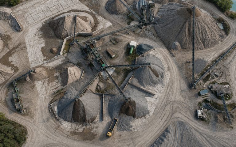 Eine Luftaufnahme zeigt ein Kieswerk mit mehreren Förderbändern, Materialhalden, Fahrwegen und Aufbereitungsanlagen. Die hellen Sand- und Kiesflächen sind klar strukturiert angeordnet.