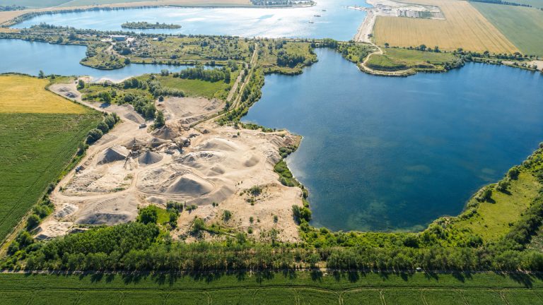 Das Bild zeigt eine weitläufige Luftaufnahme eines Kiesabbaugebiets zwischen mehreren Seen und landwirtschaftlichen Flächen. Im Vordergrund sind helle Sand- und Kieshalden, Fahrwege und Förderanlagen zu sehen, die direkt an das Ufer eines großen Gewässers grenzen. Im Hintergrund erstrecken sich weitere Seen, Grünflächen, Felder und Wege in einer offenen Landschaft.