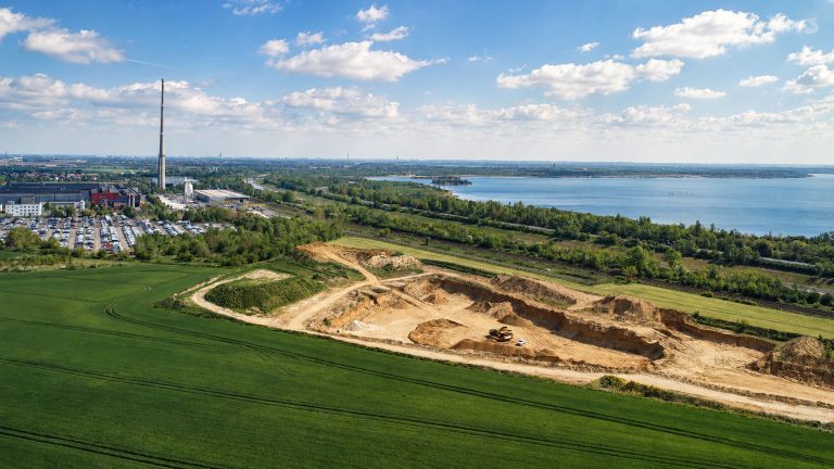 Das Bild zeigt eine Luftaufnahme eines offenen Sandabbaugebiets am Rand einer großen Wasserfläche. Im Vordergrund sind grüne Felder und die freigelegten Abbauflächen mit Fahrwegen, Erdhaufen und Arbeitsbereichen zu sehen. Im Hintergrund erstrecken sich ein See, Gehölzstreifen, Industrieanlagen und eine weite Landschaft bis zum Horizont.