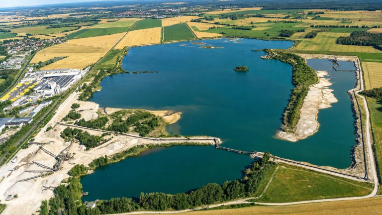 Weitläufige Luftaufnahme einer Seenlandschaft mit angrenzenden Abbauflächen, Uferzonen, Dämmen und Förderanlagen. Das Bild zeigt mehrere Wasserflächen, landwirtschaftlich genutzte Felder, Wege sowie Betriebs- und Uferbereiche aus der Vogelperspektive. Im Hintergrund erstrecken sich Ortschaften, Waldflächen und offene Landschaft bis zum Horizont.