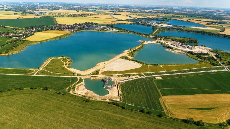 Weitläufige Luftaufnahme einer Seen- und Landschaftsfläche mit mehreren Gewässern, Feldern, Wegen und einem Kiesabbaugebiet. Im Vordergrund sind landwirtschaftlich genutzte Flächen und ein kleiner Förderbereich zu sehen, im Mittelgrund mehrere große Seen mit Uferzonen und Abbauflächen. Im Hintergrund erstrecken sich weitere Felder, Ortschaften und Verkehrswege bis zum Horizont.