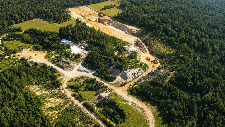 Luftaufnahme eines von Wald umgebenen Steinbruchs mit Zufahrtswegen, Abbauflächen, offenen Erd- und Gesteinsbereichen sowie kleineren Betriebsflächen. Das Bild zeigt die Anlage aus der Vogelperspektive mit dichtem Baumbestand, hellen Fahrspuren und den freigelegten Bereichen des Rohstoffabbaus.