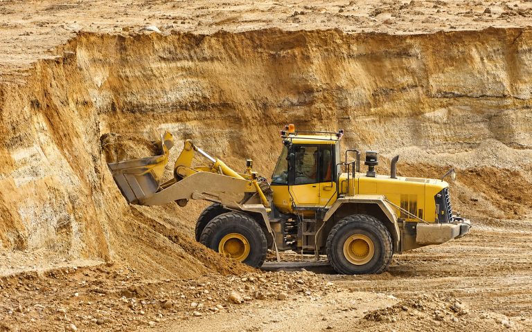 Ein gelber Radlader arbeitet an einer steilen Sandwand im Kieswerk. Die Szene zeigt geschichtete Erd- und Kieslagen, Fahrspuren und eine helle, trockene Abbaufläche.
