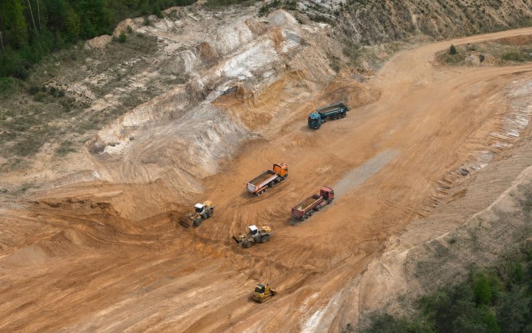 Eine Luftaufnahme zeigt ein großes Sandabbaugebiet mit orangefarbenen Abbauflächen, Fahrwegen, Böschungen und mehreren Arbeitsfahrzeugen. Die Grube ist von Waldflächen umgeben.