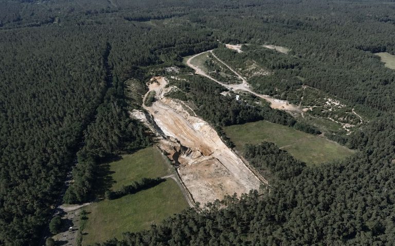 Eine hohe Luftaufnahme zeigt eine langgezogene Sandgrube, die sich durch ein großes Waldgebiet zieht. Sichtbar sind helle Abbauflächen, Zufahrtswege, Lichtungen und umgebende Grünflächen.