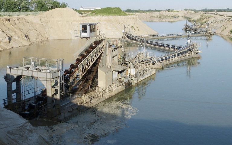 Eine industrielle Kiesgewinnungsanlage liegt auf einem ruhigen See. Förderbänder, Plattformen und sandige Ufer prägen die helle Steinbruchlandschaft.