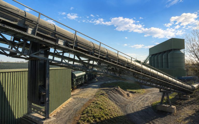 Eine große überdachte Förderbandanlage verläuft diagonal über ein industrielles Betriebsgelände. Im Hintergrund stehen grüne Silos, Hallen und weitere Anlagenteile unter klarem Himmel.