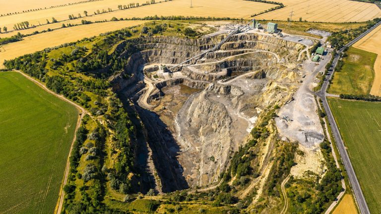Das Bild zeigt eine weitläufige Luftaufnahme eines Steinbruchs mit terrassenförmig angelegten Abbauflächen, Fahrwegen und technischen Anlagen. Der Steinbruch ist von Feldern, Straßen und Grünflächen umgeben. Im oberen Bereich der Anlage sind Fördertechnik und Betriebsflächen zu erkennen, während sich die offene Abbaugrube tief in das Gelände einschneidet.
