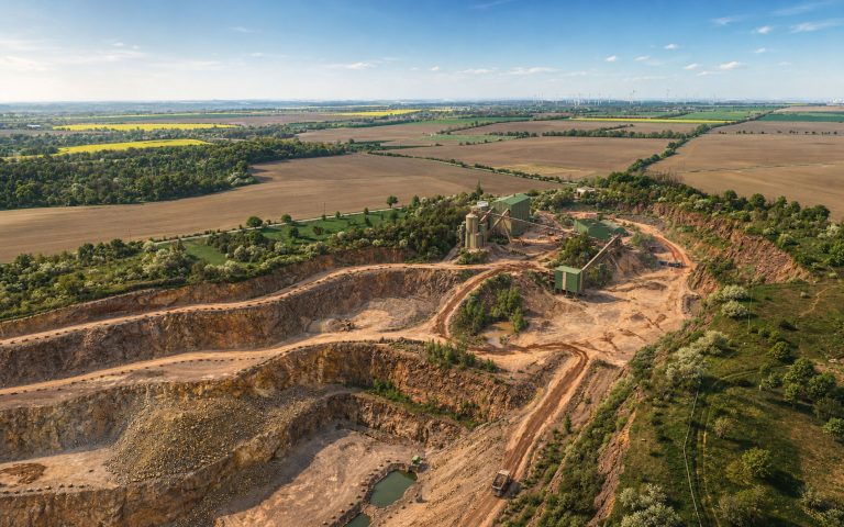 Eine Luftaufnahme zeigt einen weitläufigen Steinbruch mit terrassierten Abbauflächen, Fahrwegen und grünen Aufbereitungsanlagen. Im Hintergrund erstrecken sich Felder, Baumreihen und Windkraftanlagen.