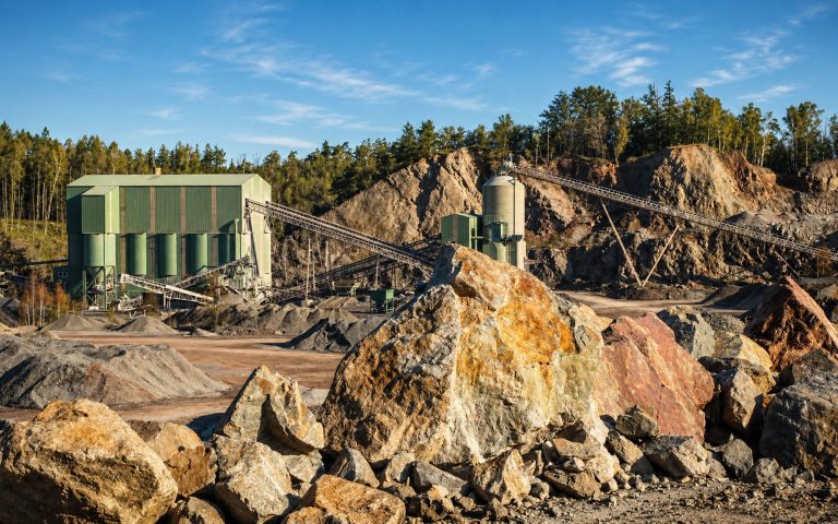 Große Gesteinsbrocken liegen im Vordergrund einer Steinbruchanlage. Dahinter sind grüne Betriebsgebäude, Silos, Förderbänder und Felswände unter hellem Himmel zu sehen.