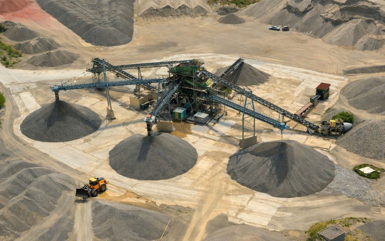 Eine Luftaufnahme zeigt ein Kieswerk mit zentraler Förder- und Sortieranlage, mehreren Förderbändern sowie großen Materialhalden auf dem Werksgelände.