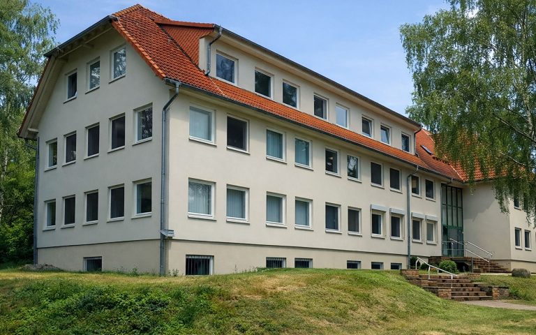 Das Verwaltungsgebäude der MDB am Hauptsitz Sennewitz mit heller Fassade, rotem Ziegeldach und mehreren Fensterreihen. Das Gebäude liegt in einer grünen Umgebung mit Bäumen, Rasenfläche und Treppenaufgang.